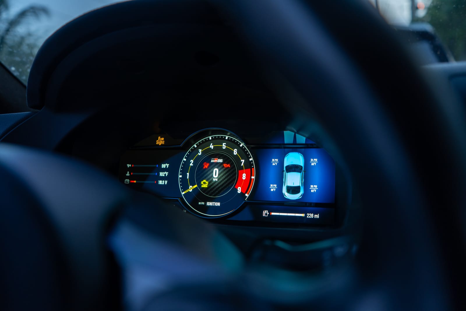 The GRZ Cluster fitted to a Toyota GR86, seen through the steering wheel at dusk: oil and coolant temperatures on the left, the tachometer in the centre, and per-corner tire pressures on the 3D car to the right